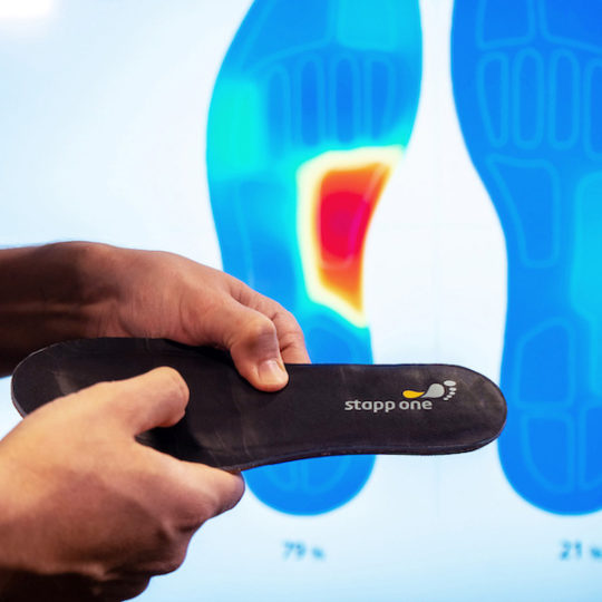 Thumb presses on sensor sole and screen in background shows live biofeedback of pressure
